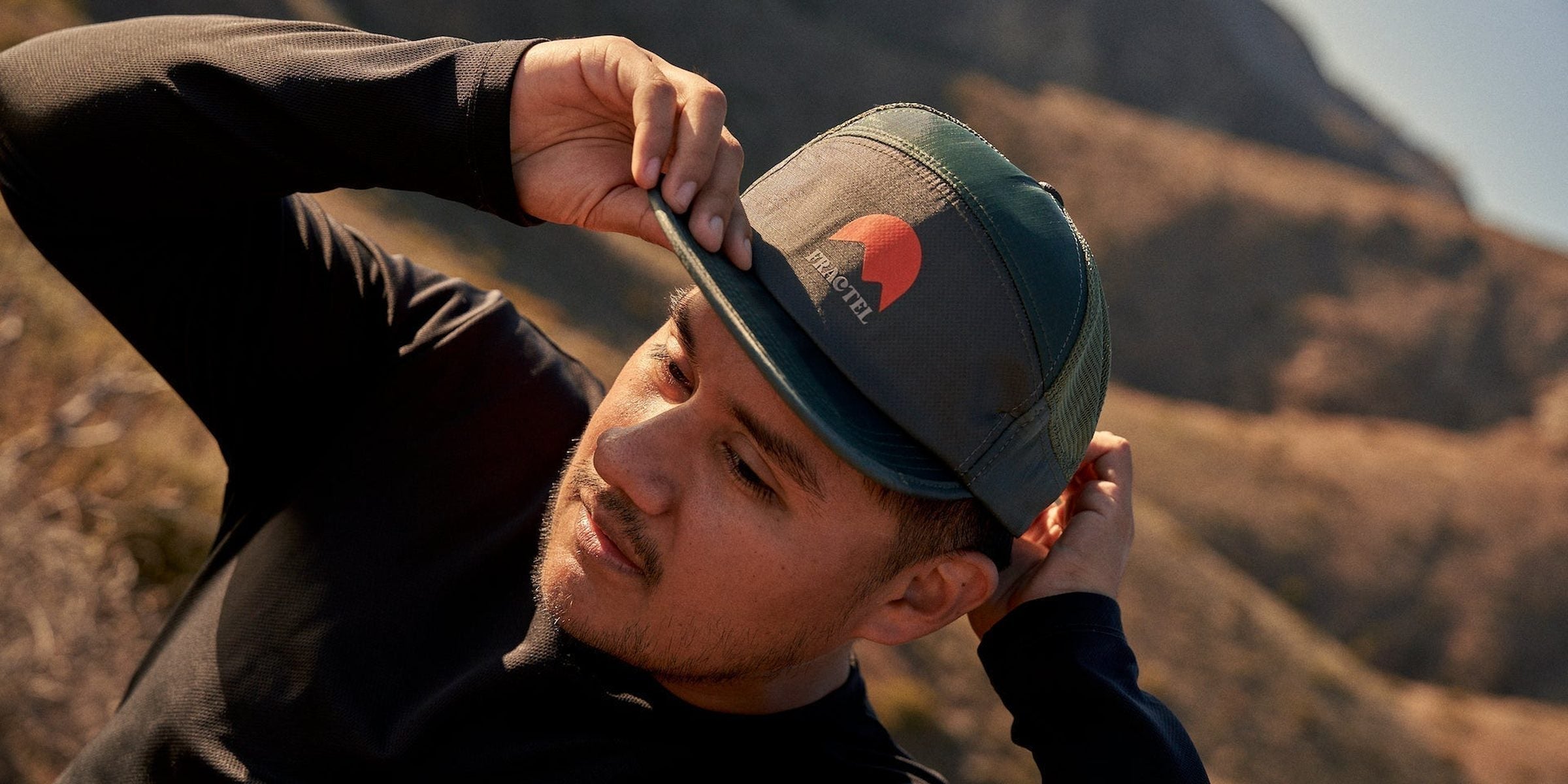 Man adjusting a dark green Fractel running trucker hat with a sunrise logo on a hillside