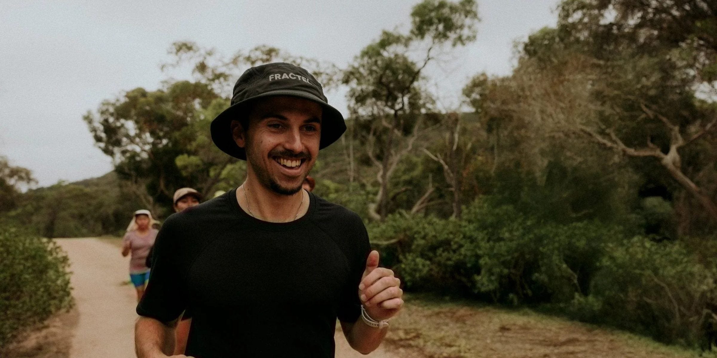 Smiling runner in a black Fractel running bucket hat and tee on a bush trail with other runners behind him