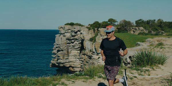 Runner in a multicoloured Fractel cap, black tee and printed shorts on a coastal cliff path, new running hats and gear