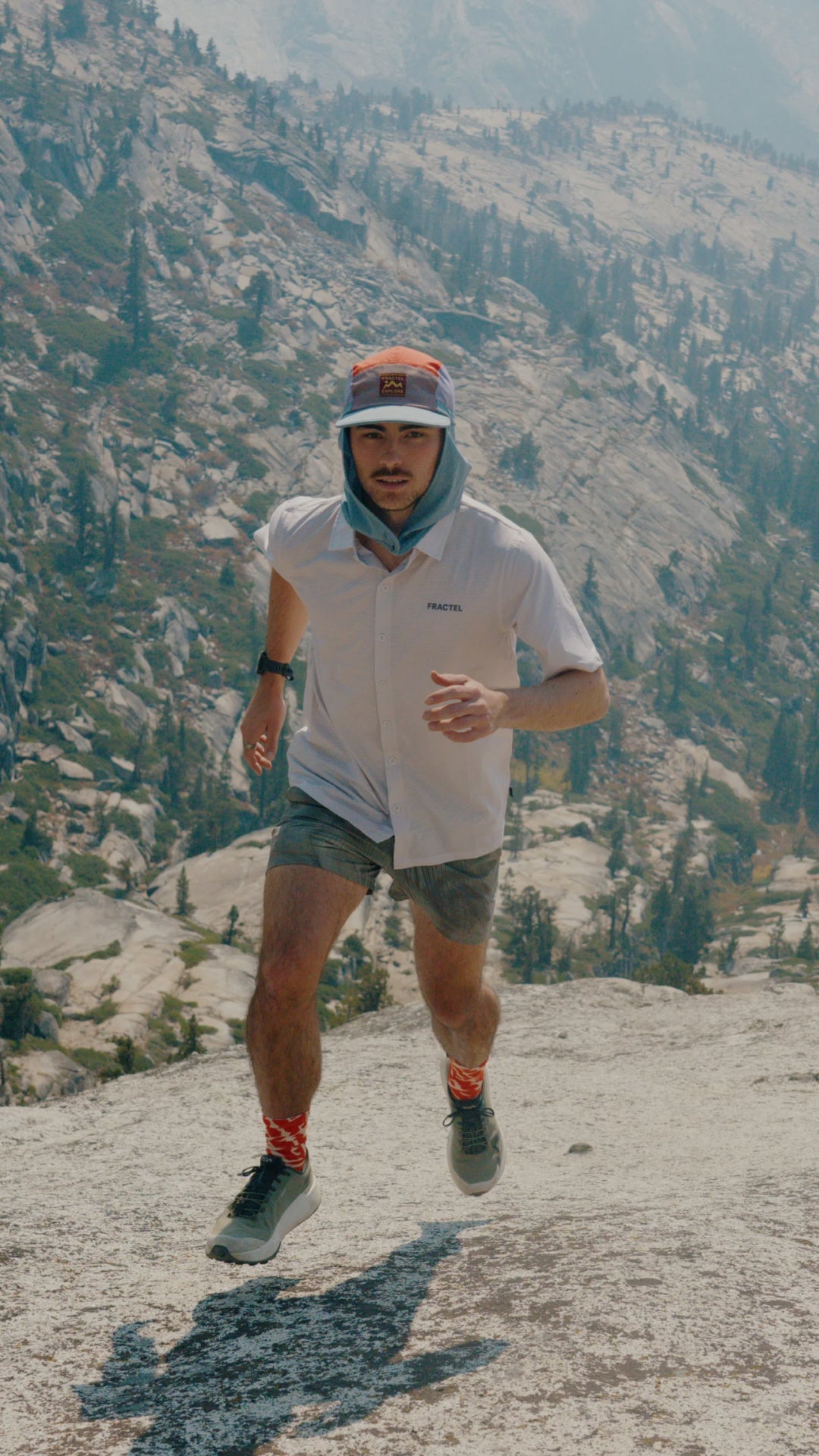 A male trail runner is captured mid-stride on a rocky mountain path, wearing a white short-sleeve performance shirt, gray shorts, and a white cap with a red-orange logo. The background shows a vast, sunlit mountain landscape.