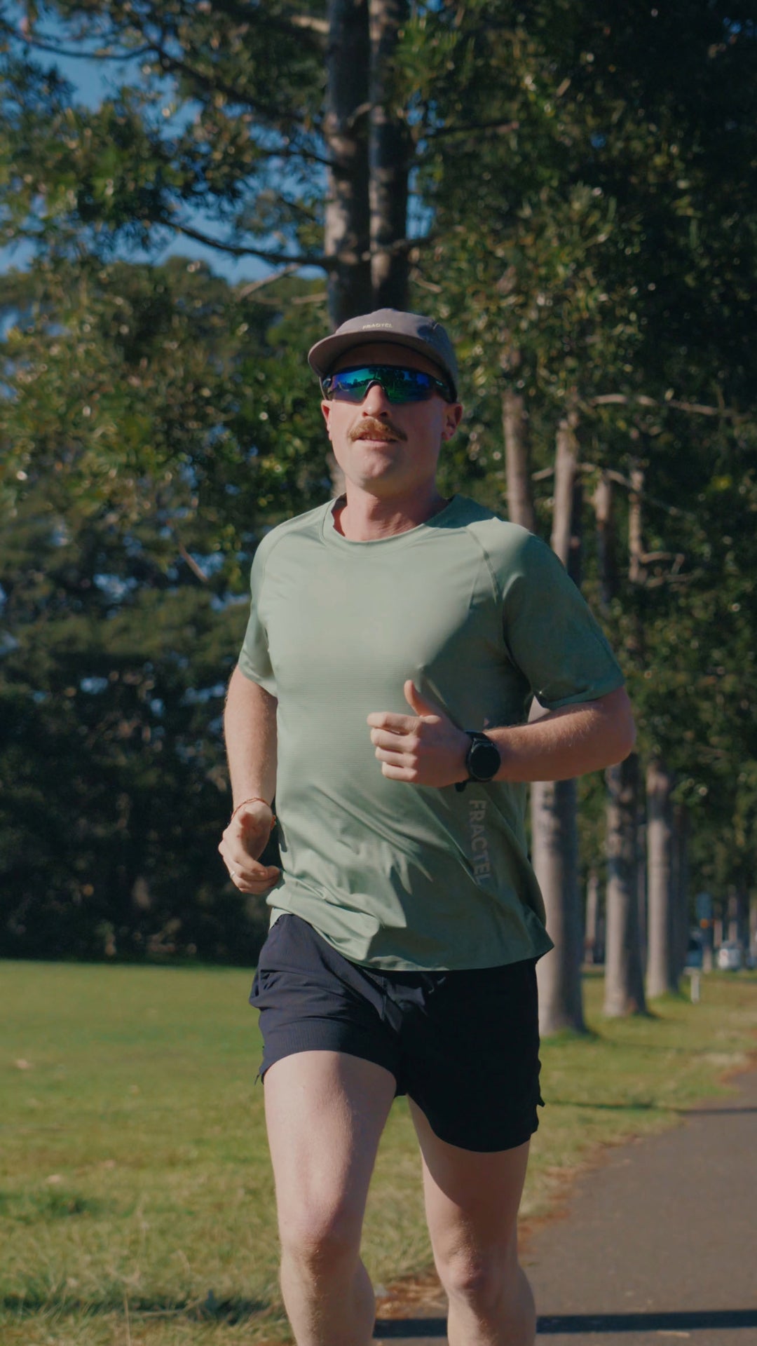 A man wearing a light green t-shirt, dark shorts, and a cap is jogging on a tree-lined path outdoors.