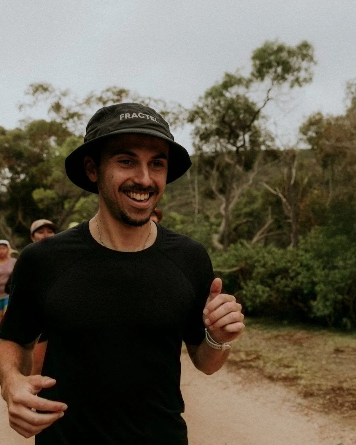 Runner in a black Fractel bucket hat smiling on a dirt bush trail